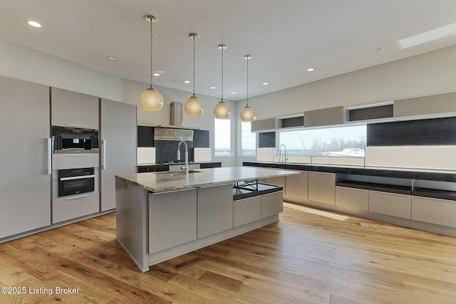 kitchen with decorative light fixtures, black oven, light wood finished floors, and an island with sink