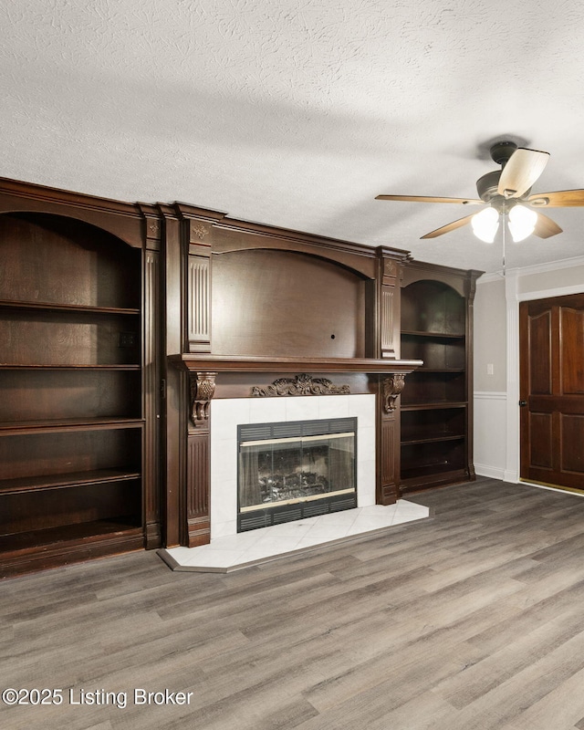 unfurnished living room featuring a textured ceiling, ceiling fan, wood finished floors, a tiled fireplace, and crown molding