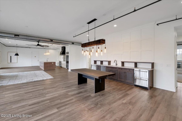 kitchen with wood finished floors, a ceiling fan, hanging light fixtures, light countertops, and wall chimney exhaust hood