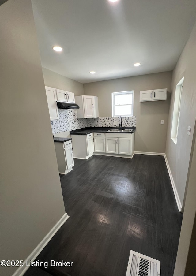 kitchen with dark countertops, backsplash, white cabinetry, a sink, and under cabinet range hood