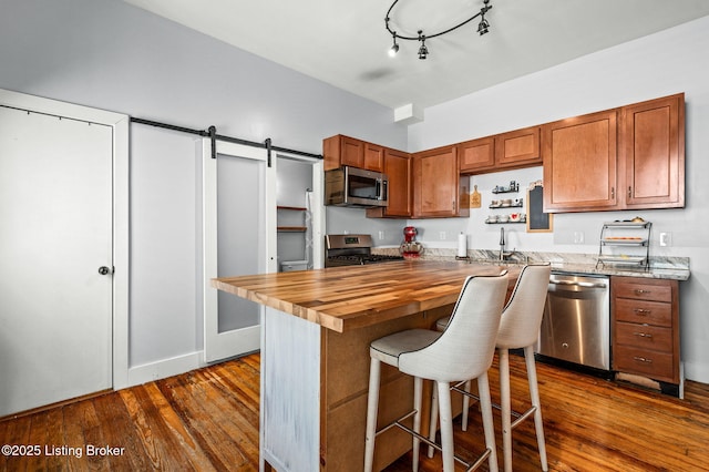 kitchen with a barn door, dark wood finished floors, appliances with stainless steel finishes, brown cabinets, and wooden counters