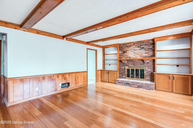 unfurnished living room with built in features, a wainscoted wall, light wood-style floors, a fireplace, and beam ceiling