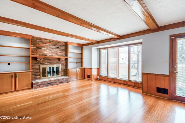 unfurnished living room featuring a wainscoted wall, light wood-style flooring, beamed ceiling, a textured ceiling, and a fireplace