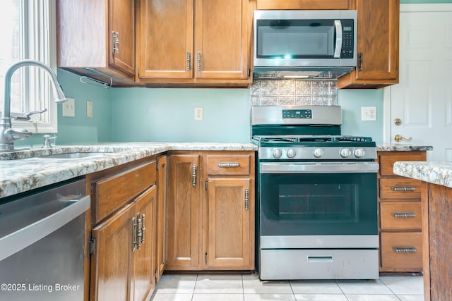 kitchen featuring light stone counters, light tile patterned floors, stainless steel appliances, brown cabinetry, and a sink