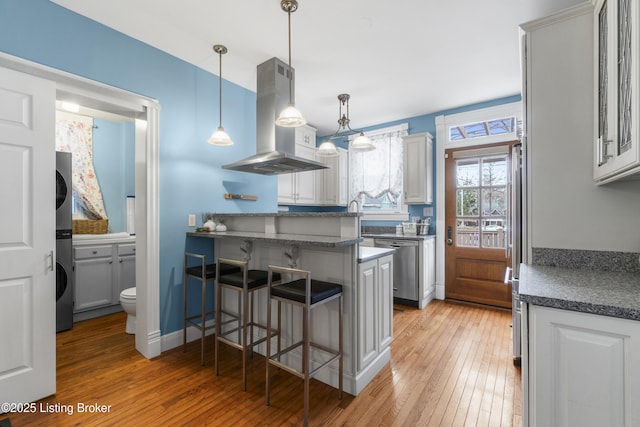 kitchen featuring dark countertops, hanging light fixtures, stainless steel dishwasher, island range hood, and a kitchen bar