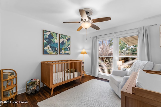 bedroom featuring a crib, ceiling fan, baseboards, and dark wood-style flooring