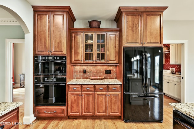 kitchen with arched walkways, light wood-type flooring, light stone countertops, black appliances, and glass insert cabinets