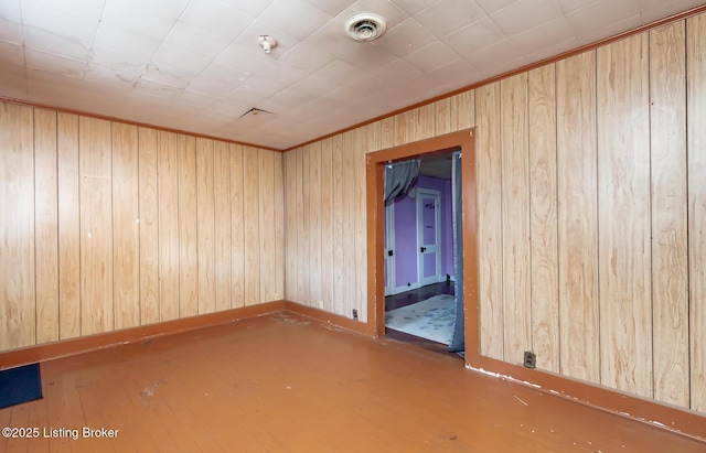 empty room with wooden walls, wood finished floors, visible vents, baseboards, and ornamental molding