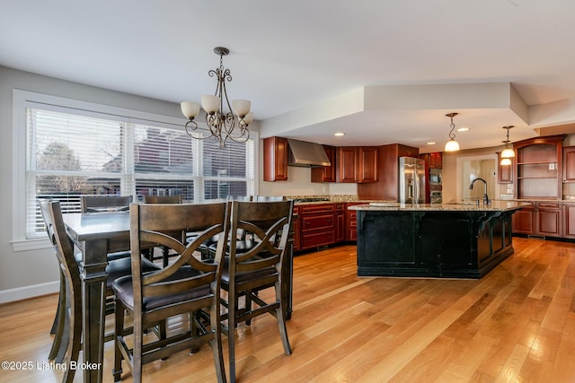 kitchen with open shelves, hanging light fixtures, appliances with stainless steel finishes, wall chimney range hood, and an island with sink