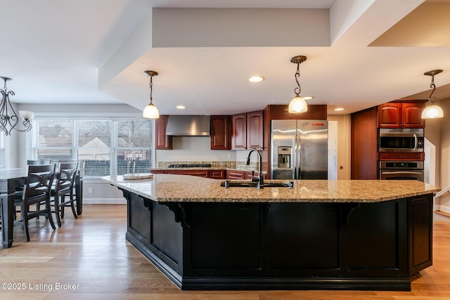 kitchen featuring wall chimney range hood, a center island with sink, stainless steel appliances, and a sink