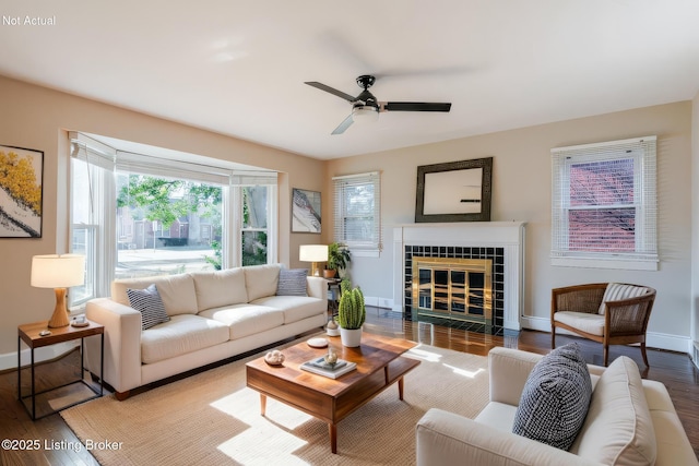 living area with ceiling fan, a tiled fireplace, wood finished floors, and baseboards