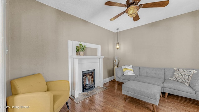 living area featuring a fireplace with flush hearth, a ceiling fan, baseboards, and wood finished floors