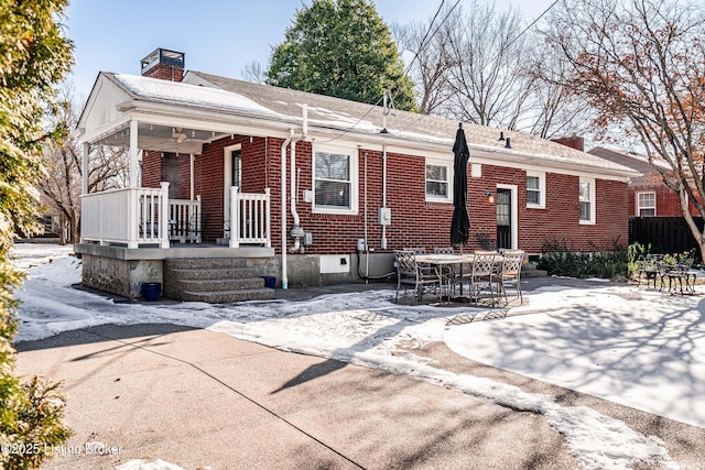 snow covered property with covered porch, a patio, brick siding, and a chimney