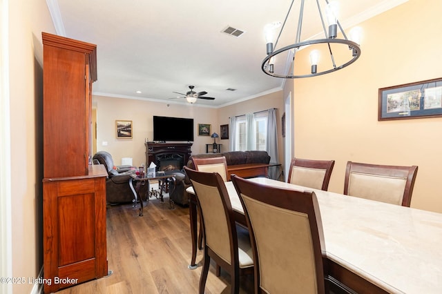 dining area with light wood-style floors, ceiling fan with notable chandelier, crown molding, and a glass covered fireplace