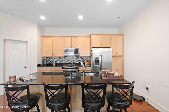 kitchen with light brown cabinets, stainless steel appliances, backsplash, dark countertops, and a center island with sink