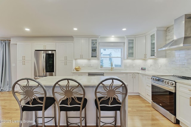 kitchen featuring stainless steel appliances, wall chimney range hood, a kitchen bar, and light wood-style flooring