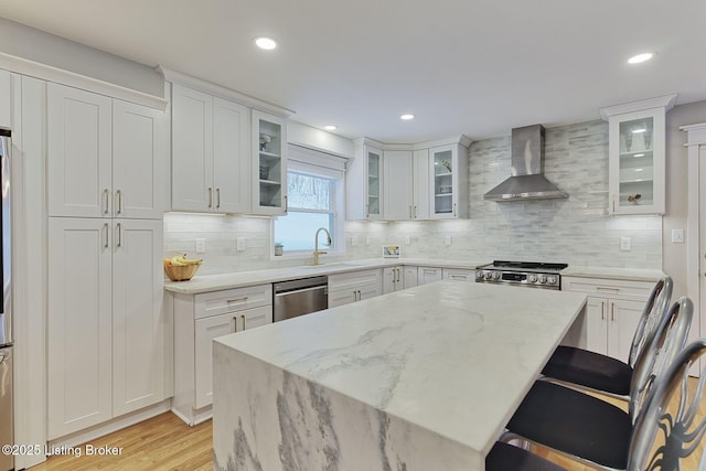 kitchen with light wood finished floors, appliances with stainless steel finishes, light stone counters, wall chimney range hood, and white cabinetry