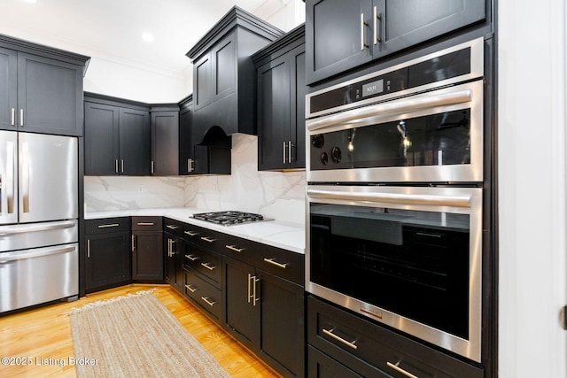 kitchen featuring stainless steel appliances, ornamental molding, light wood-type flooring, backsplash, and light stone countertops