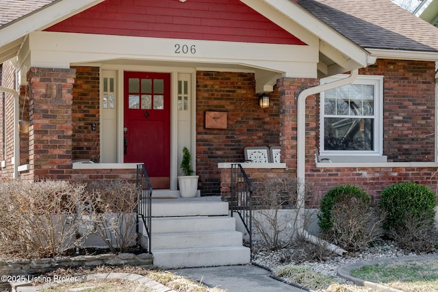 doorway to property featuring a shingled roof, covered porch, and brick siding