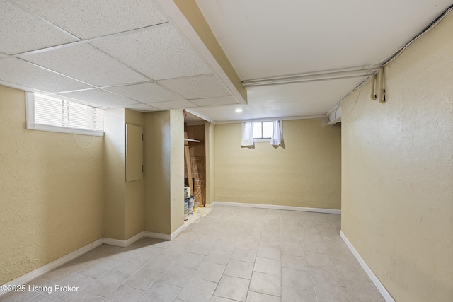 basement with a textured wall, baseboards, a drop ceiling, and electric panel