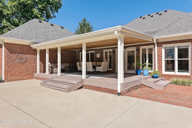 back of house with brick siding, a shingled roof, an outdoor living space, and a patio