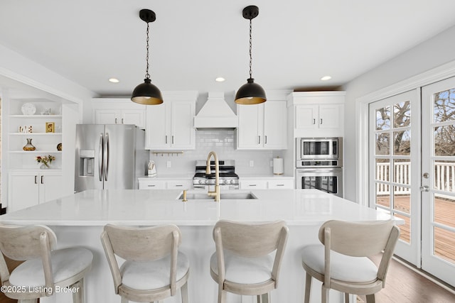 kitchen with stainless steel appliances, light countertops, an island with sink, decorative light fixtures, and custom range hood