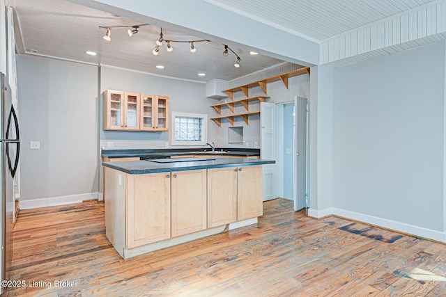 kitchen featuring light wood finished floors, dark countertops, glass insert cabinets, light brown cabinets, and baseboards