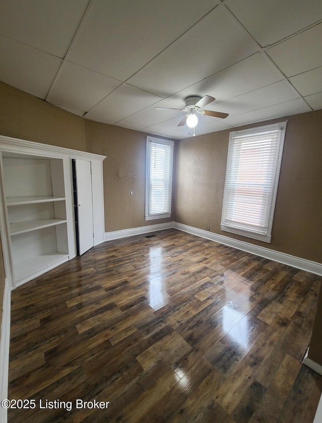 unfurnished bedroom featuring a ceiling fan, baseboards, dark wood-style flooring, a drop ceiling, and a closet