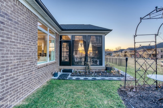 exterior entry at dusk featuring brick siding, a lawn, a shingled roof, and fence