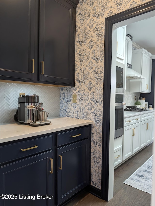 kitchen with wallpapered walls, stainless steel appliances, light countertops, white cabinetry, and wall chimney exhaust hood