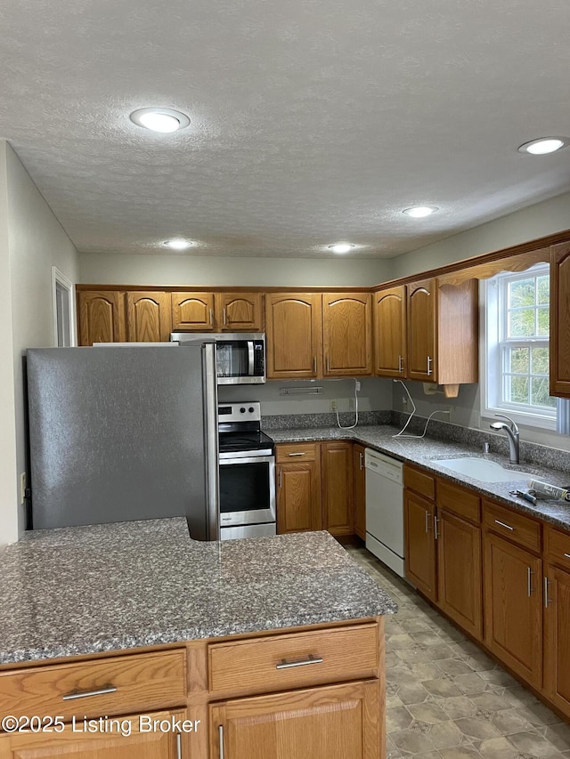 kitchen featuring a textured ceiling, a sink, appliances with stainless steel finishes, brown cabinets, and dark stone counters