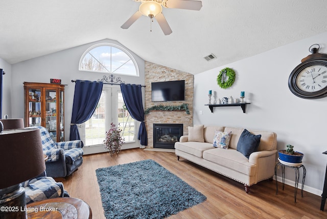 living room featuring ceiling fan, a stone fireplace, wood finished floors, visible vents, and vaulted ceiling