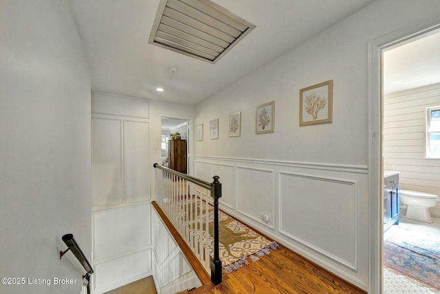 hall with a wainscoted wall, a decorative wall, wood finished floors, and an upstairs landing