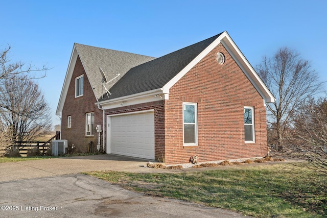 view of side of property with driveway, roof with shingles, a garage, central air condition unit, and brick siding