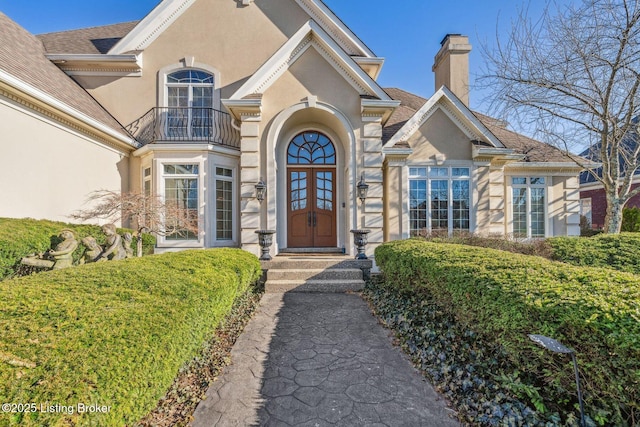 view of exterior entry with french doors, a chimney, a balcony, and stucco siding