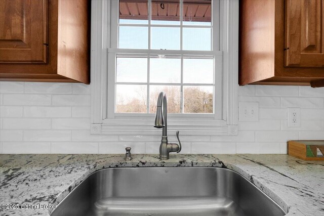 interior details featuring brown cabinetry, a sink, decorative backsplash, and light stone countertops