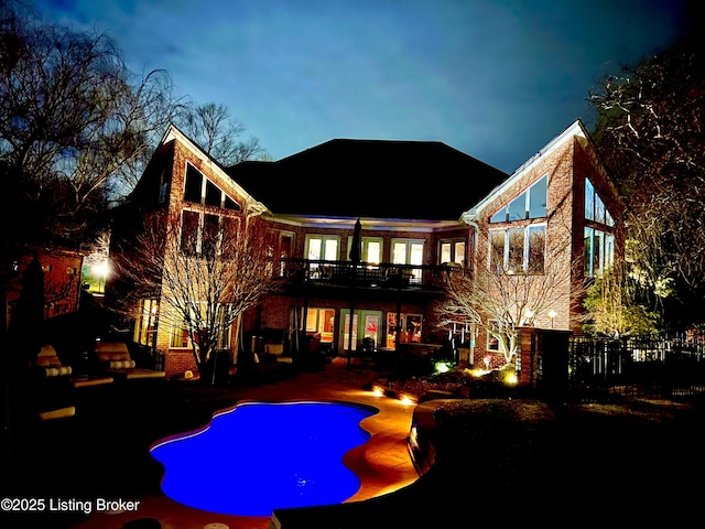 back of house at twilight featuring fence, an outdoor pool, a deck, a patio area, and brick siding