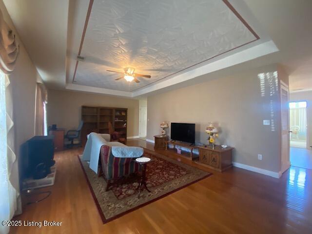 living area featuring a tray ceiling, wood finished floors, a ceiling fan, and baseboards