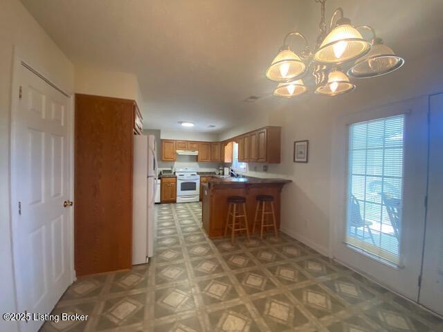 kitchen with a breakfast bar area, an inviting chandelier, brown cabinetry, white appliances, and a peninsula