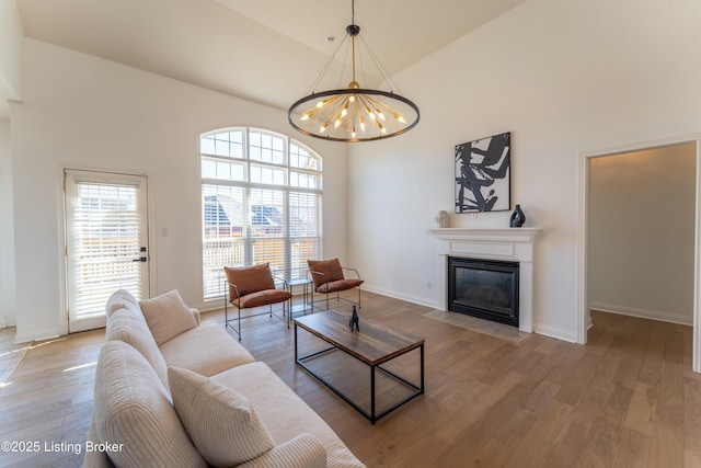 living room with a notable chandelier, a high ceiling, a fireplace with flush hearth, light wood-type flooring, and baseboards