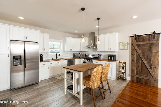 kitchen with wooden counters, stainless steel appliances, light wood-style floors, a barn door, and wall chimney exhaust hood