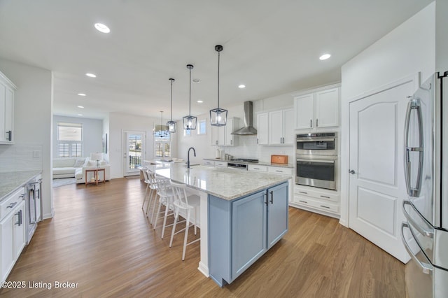 kitchen with a sink, wood finished floors, white cabinets, appliances with stainless steel finishes, and wall chimney exhaust hood