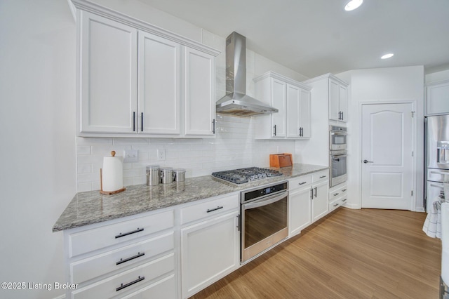 kitchen with stainless steel appliances, light wood-style floors, white cabinets, wall chimney exhaust hood, and tasteful backsplash