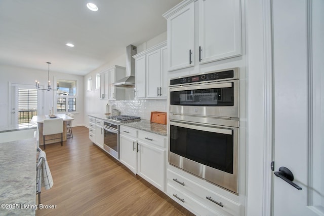 kitchen with tasteful backsplash, appliances with stainless steel finishes, white cabinetry, light wood-type flooring, and wall chimney exhaust hood