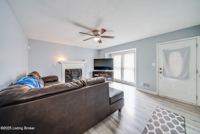 living area featuring light wood finished floors, visible vents, a glass covered fireplace, ceiling fan, and baseboards