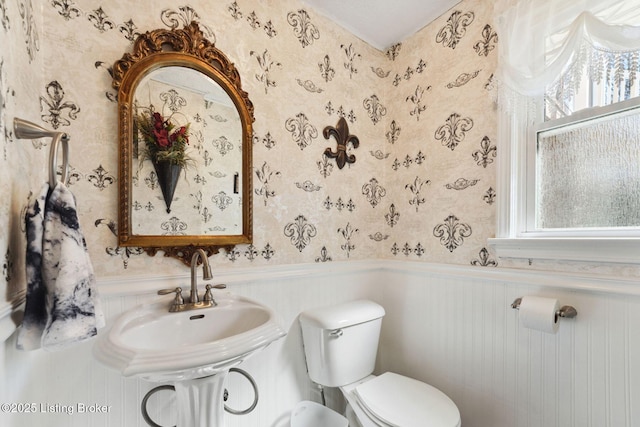 half bathroom featuring toilet, a wainscoted wall, a sink, and wallpapered walls