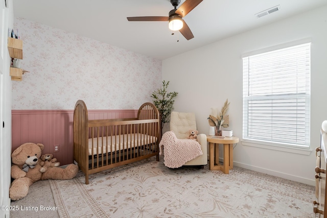 bedroom with visible vents, wainscoting, ceiling fan, a crib, and wallpapered walls