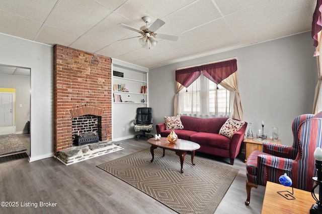 living area with baseboards, a drop ceiling, ceiling fan, wood finished floors, and a brick fireplace