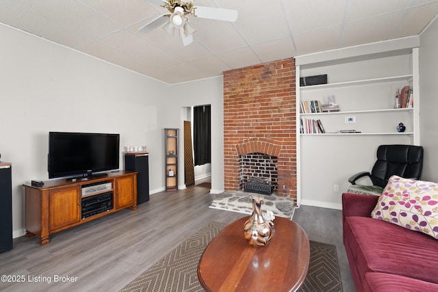 living room featuring a fireplace, ceiling fan, wood finished floors, a drop ceiling, and baseboards