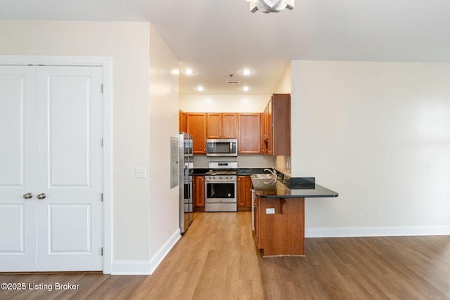 kitchen featuring stainless steel appliances, a peninsula, light wood-style floors, brown cabinets, and dark countertops